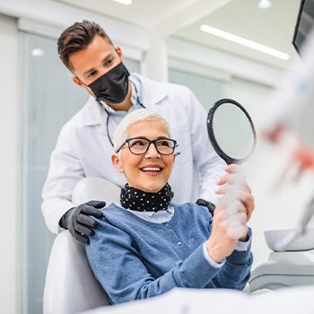 Woman with short white hair smiling at reflection with dentist touching her shoulders