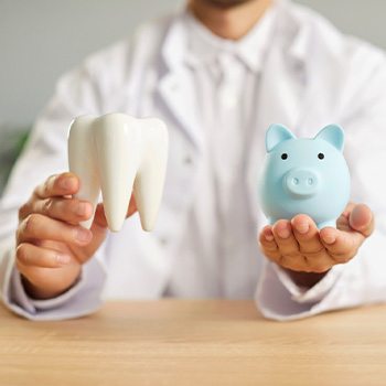 Dentist in white coat holding model tooth and blue piggy bank